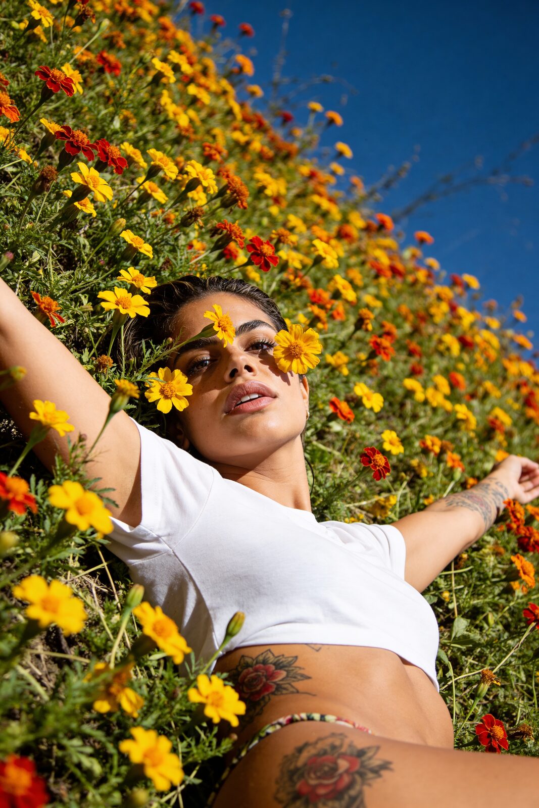 Thrown back into the flowers, sun on the ink, nothing to prove. She grew up in a culture where the body is celebration, not transgression — she knows hers, tends it, uses it as an instrument. The eroticism reads as natural because to her it is. The same body that tattoos all day dances all night; they were never two different things.